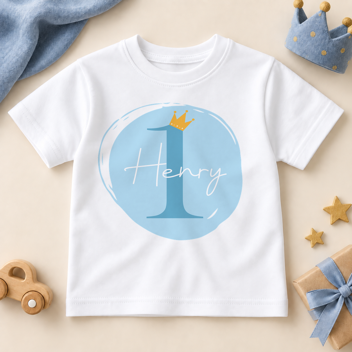 Personalised white first birthday T-shirt with a blue number one, golden crown and Henry example name in white script, styled with a blue crown, wooden car and birthday gift.