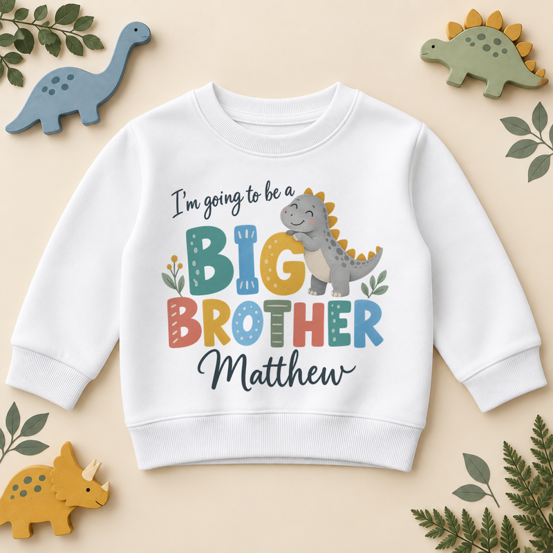 White personalised big brother sweatshirt with a cute dino design: a smiling grey dinosaur with mustard spikes hugs the G of I'm going to be a BIG BROTHER in colourful letters, with Matthew as an example name in navy script