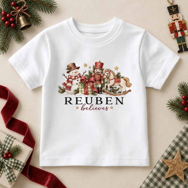 White kids T-shirt with a snowman, nutcracker, rocking horse and ribboned gift. Reuben believes example personalisation.