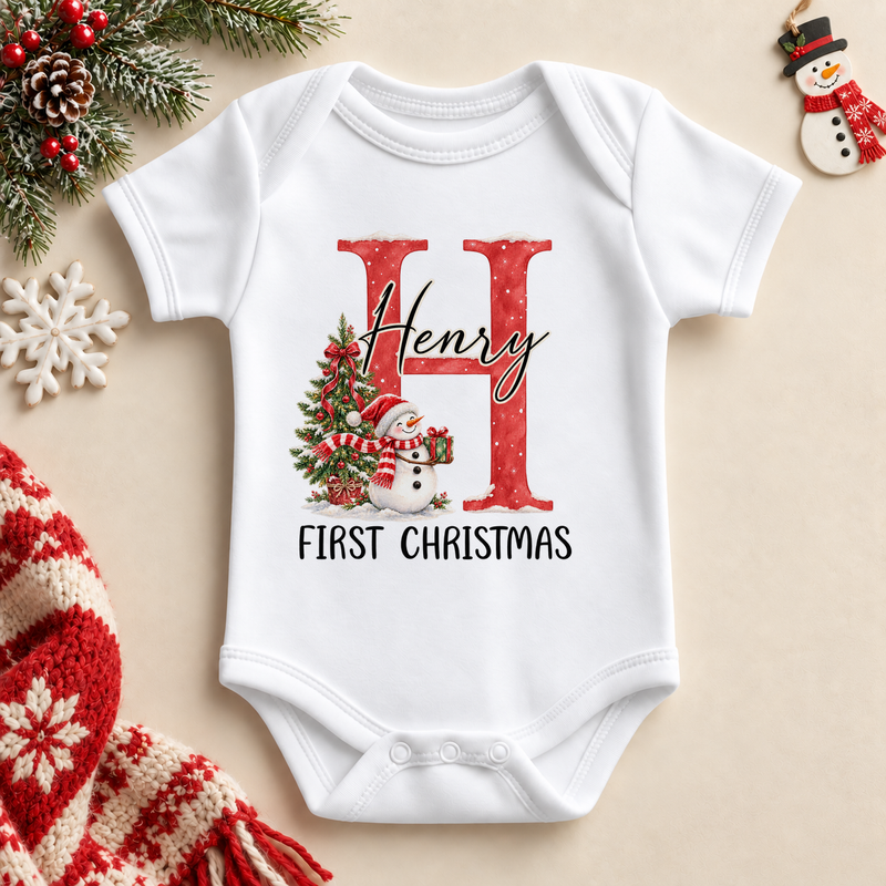 White baby bodysuit. The print features a red initial beside a snowman and a decorated Christmas tree.