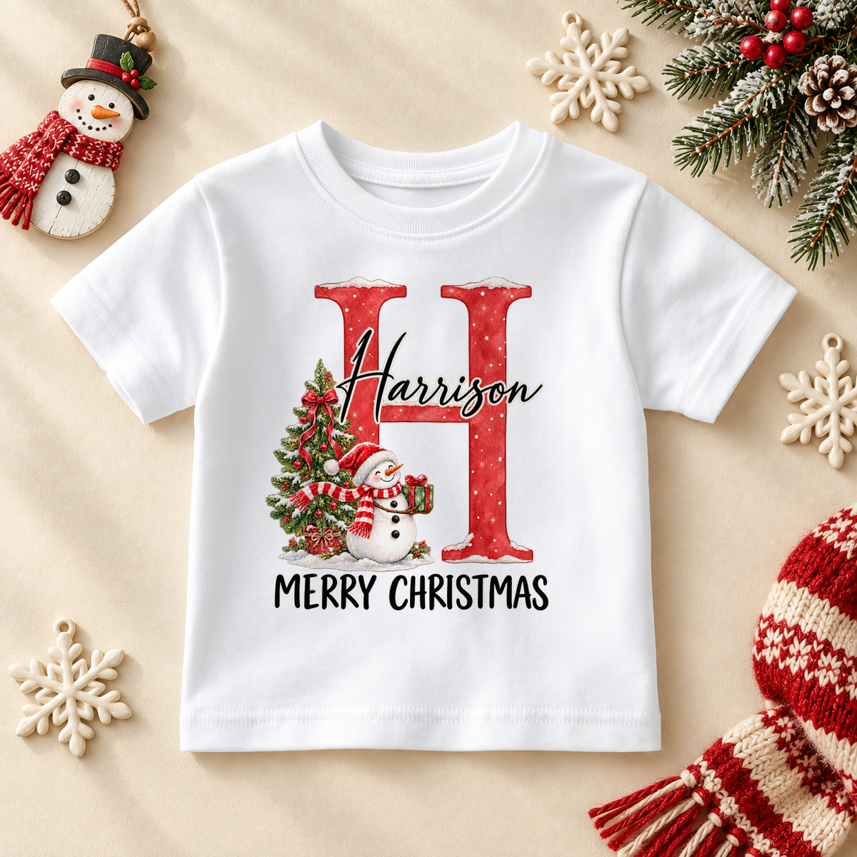 White children’s T-shirt. The print features a red initial beside a snowman and a decorated Christmas tree.