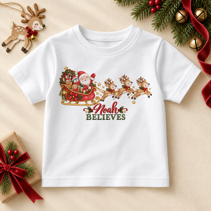White children's T-shirt with Santa's sleigh and reindeer, the example name Noah in red script and green Believes lettering.