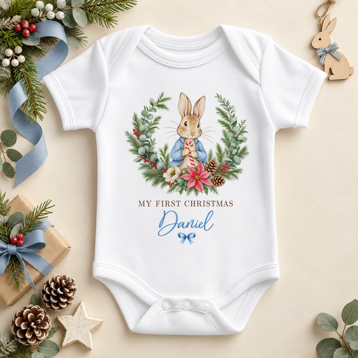 White baby bodysuit with a blue-coated bunny framed by a festive wreath with flowers and berries. My First Christmas wording. Example personalisation shown.