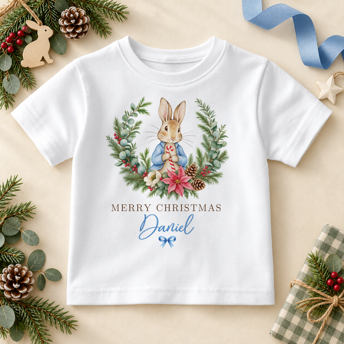 White kids T-shirt with a blue-coated bunny framed by a festive wreath with flowers and berries. Merry Christmas wording. Example personalisation shown.
