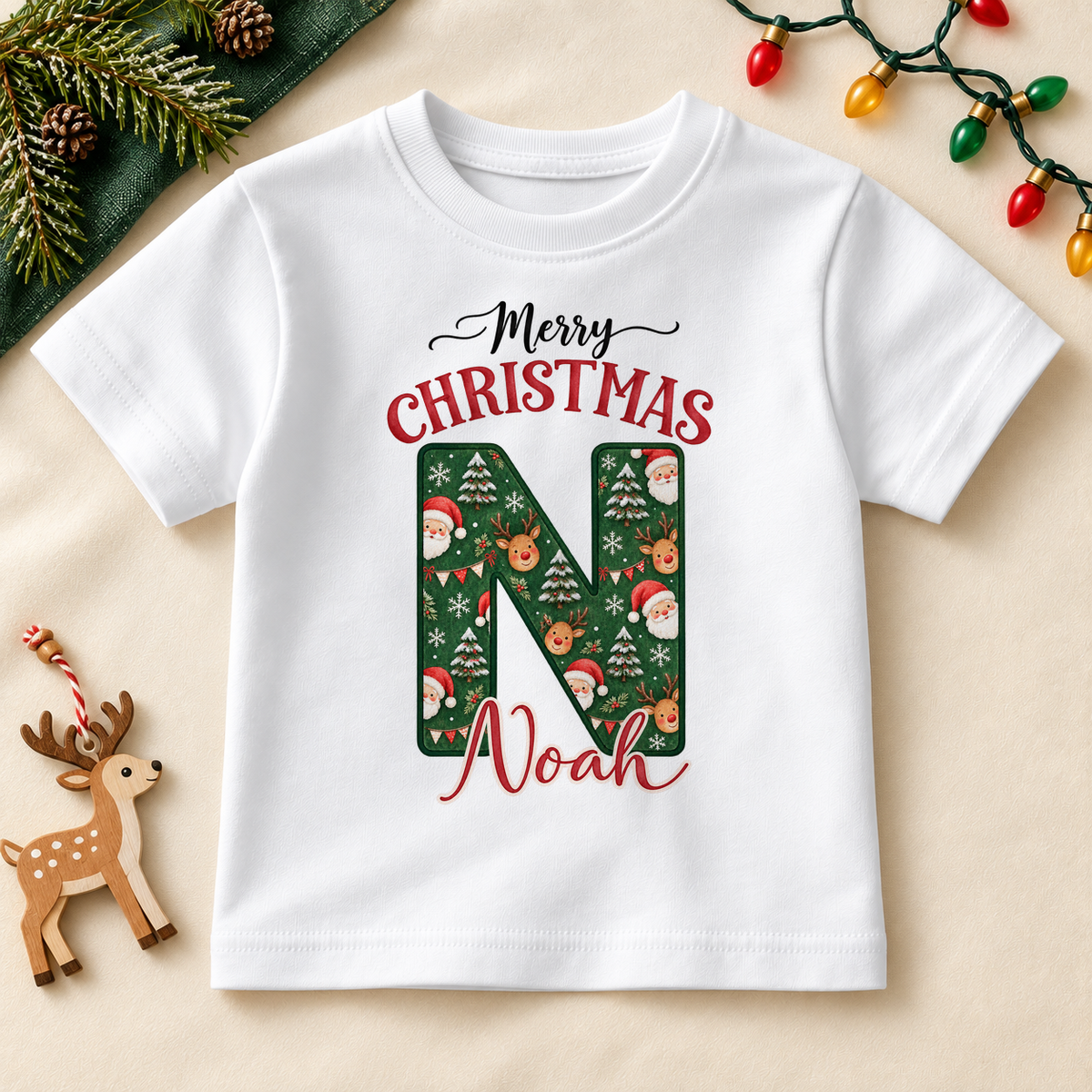 White children’s T-shirt. The print features a deep green initial filled with Santa, reindeer and snowy Christmas details.