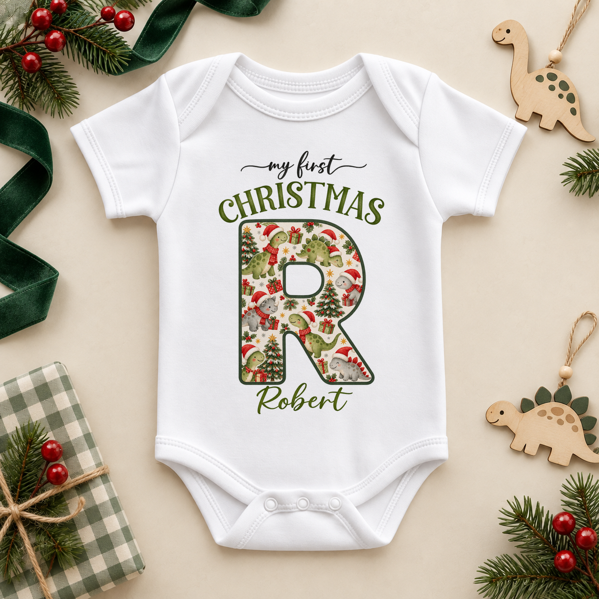 White baby bodysuit. The print features a cream initial filled with little dinosaurs in festive hats and scarves.