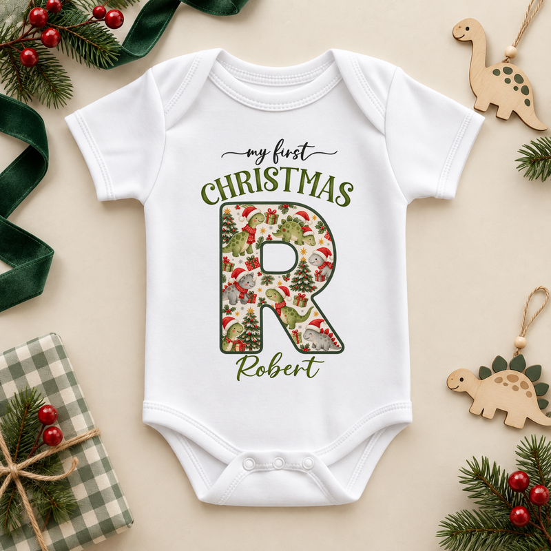 White baby bodysuit. The print features a cream initial filled with little dinosaurs in festive hats and scarves.