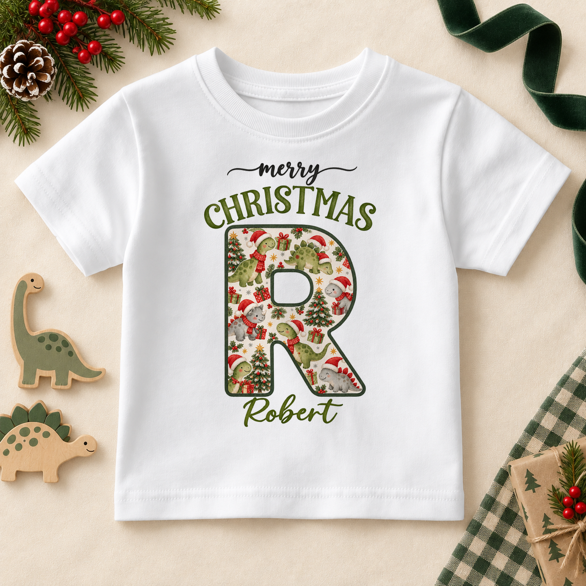 White children’s T-shirt. The print features a cream initial filled with little dinosaurs in festive hats and scarves.