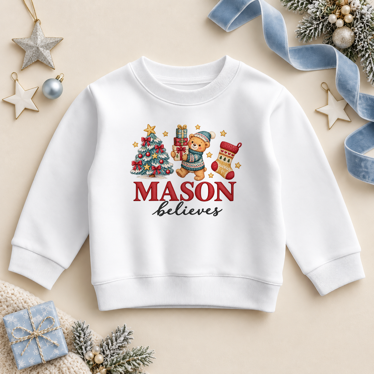 White children’s sweatshirt. The print features a teddy in a blue hat holding gifts beside a decorated tree and Christmas stocking.