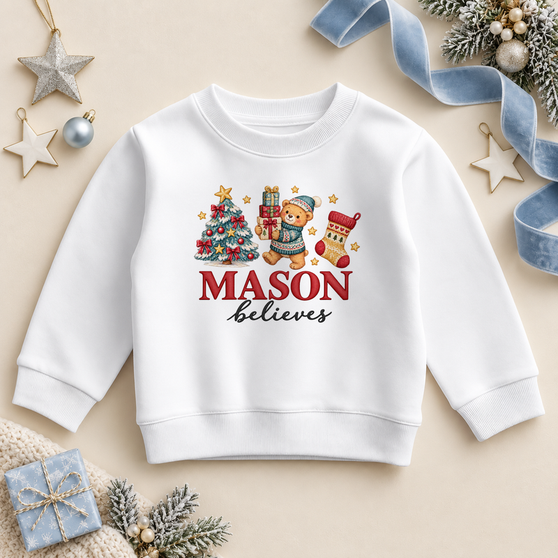 White children’s sweatshirt. The print features a teddy in a blue hat holding gifts beside a decorated tree and Christmas stocking.