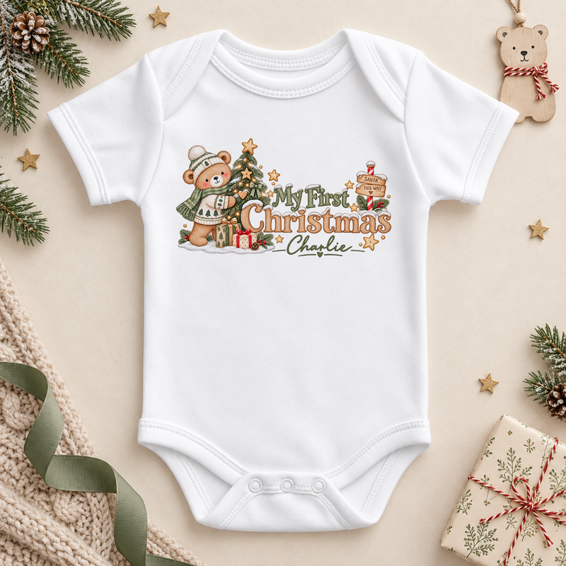 White baby bodysuit. The print features a teddy in a knitted hat and scarf beside a Christmas tree and gifts.