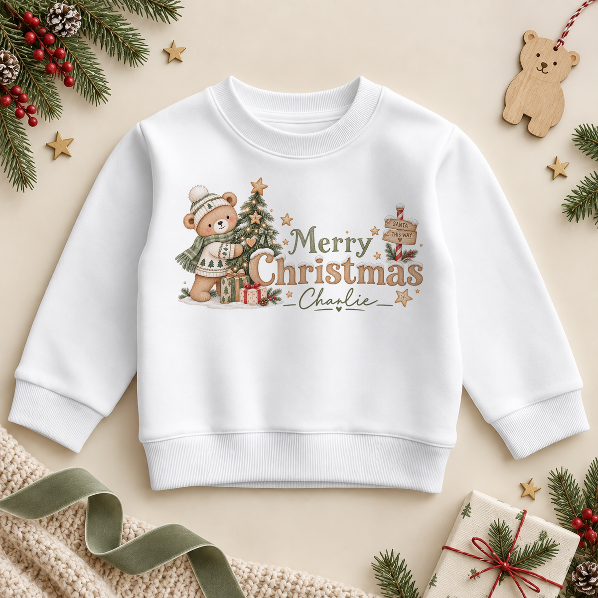White children’s sweatshirt. The print features a teddy in a knitted hat and scarf beside a Christmas tree and gifts.
