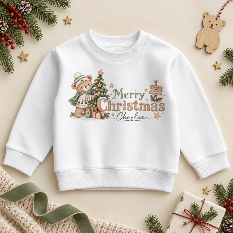 White children’s sweatshirt. The print features a teddy in a knitted hat and scarf beside a Christmas tree and gifts.