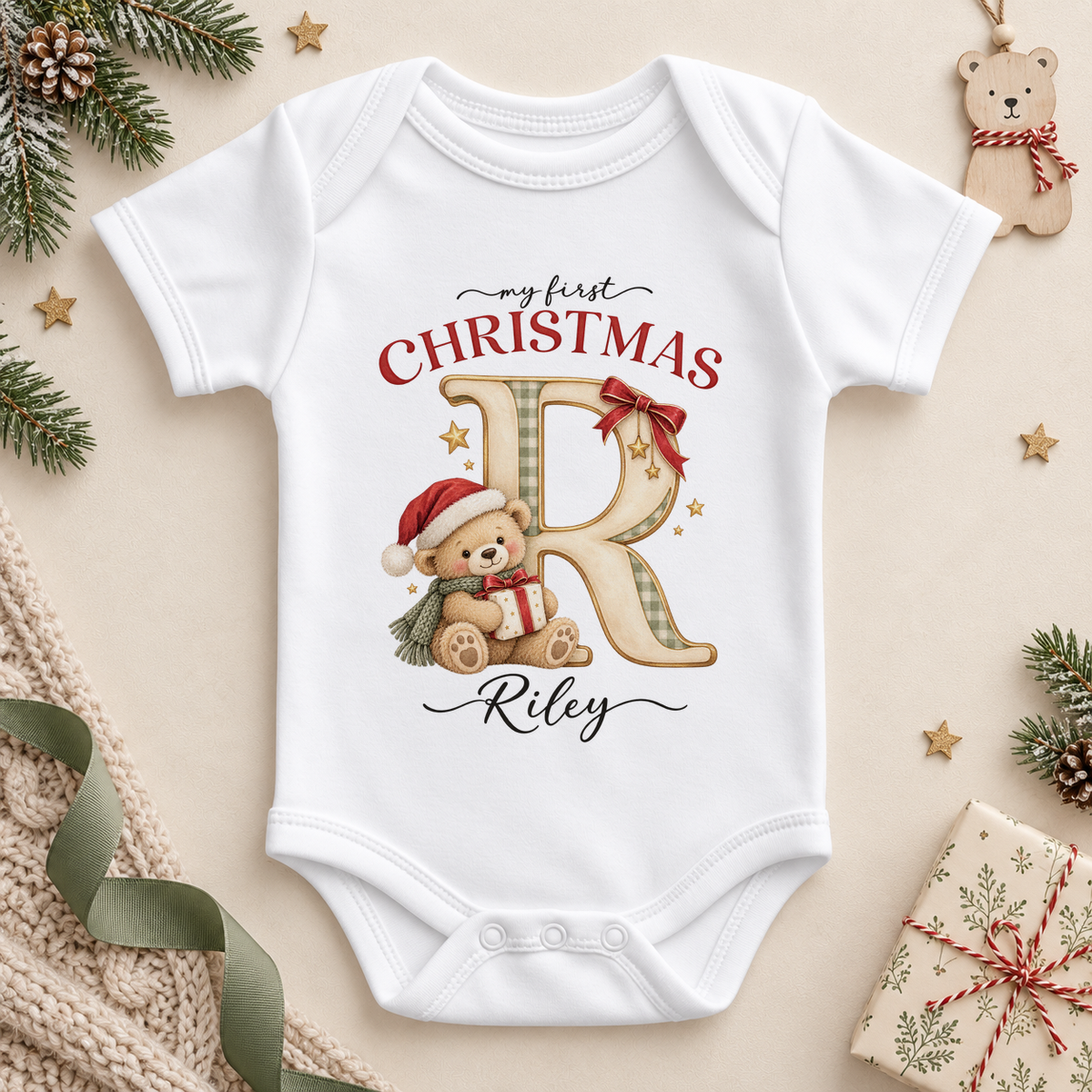 White baby bodysuit. The print features a cream and gold initial beside a teddy in a Santa hat holding a small present.