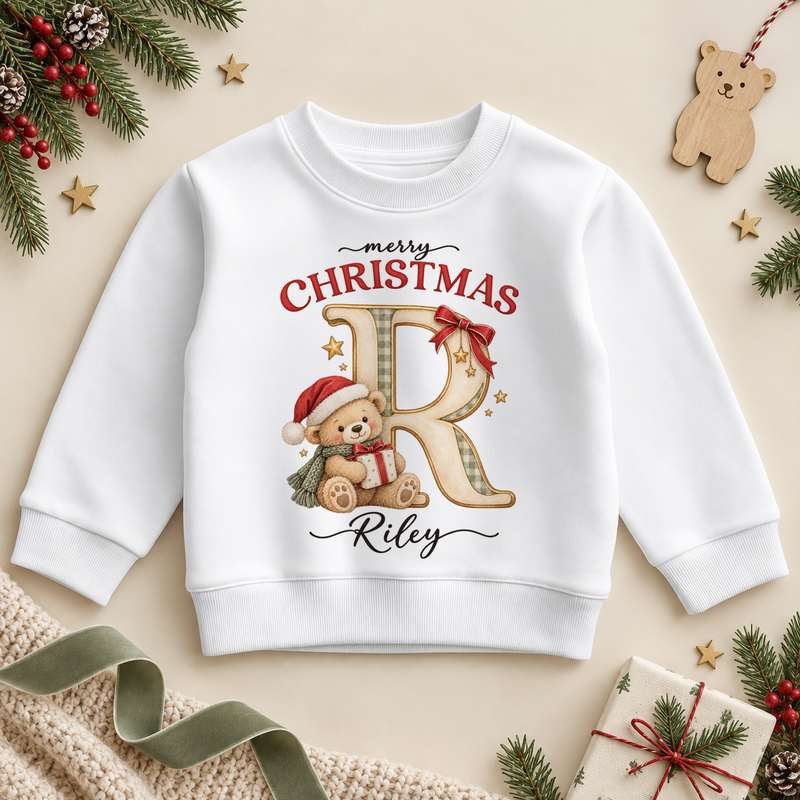 White children’s sweatshirt. The print features a cream and gold initial beside a teddy in a Santa hat holding a small present.