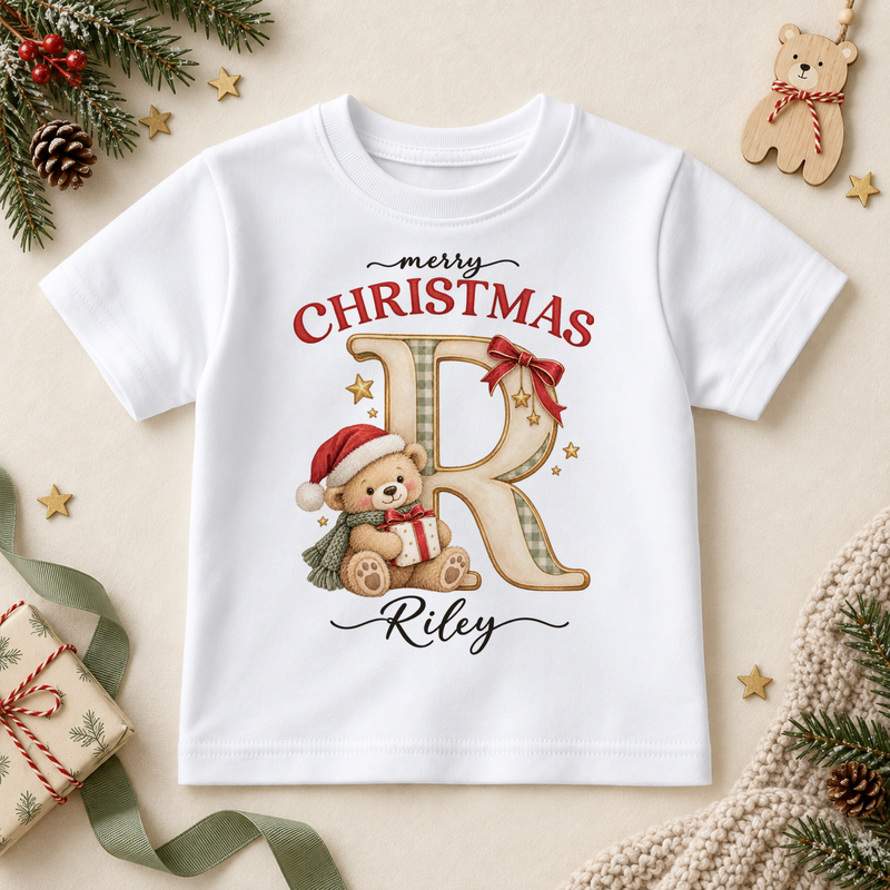 White children’s T-shirt. The print features a cream and gold initial beside a teddy in a Santa hat holding a small present.