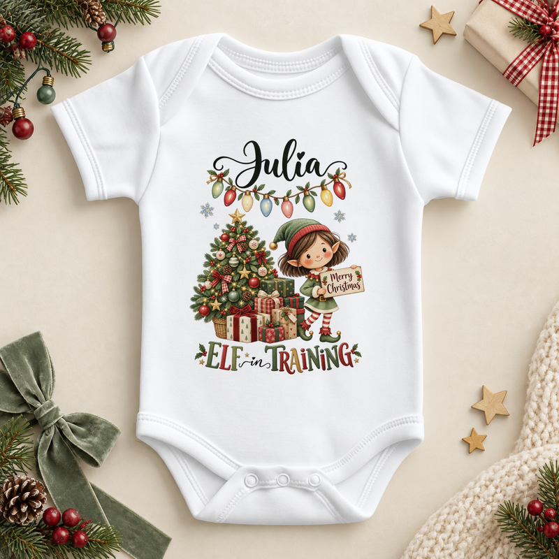 White baby bodysuit. The print features a girl elf holding a festive sign beside a tree, presents and Christmas lights.