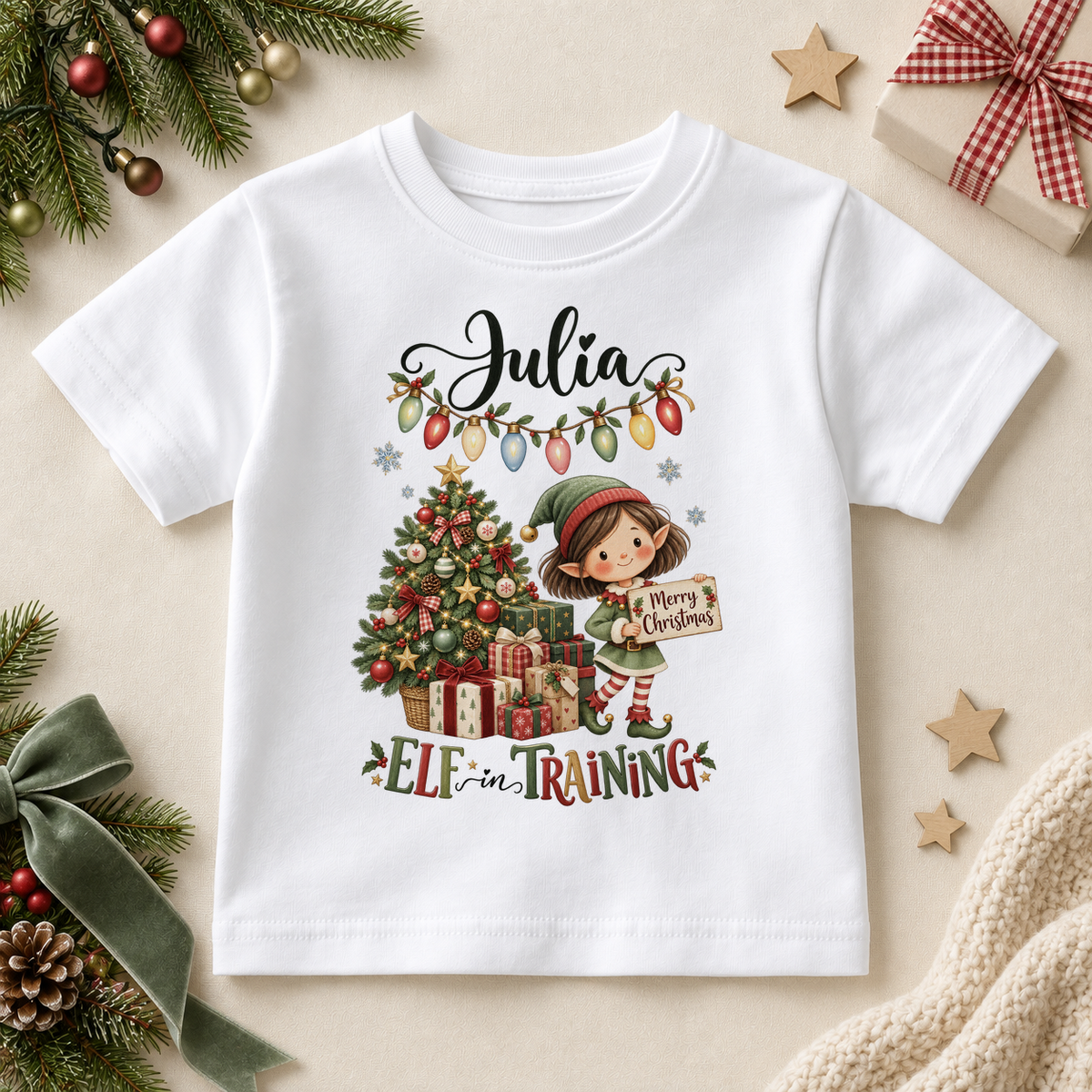 White children’s T-shirt. The print features a girl elf holding a festive sign beside a tree, presents and Christmas lights.