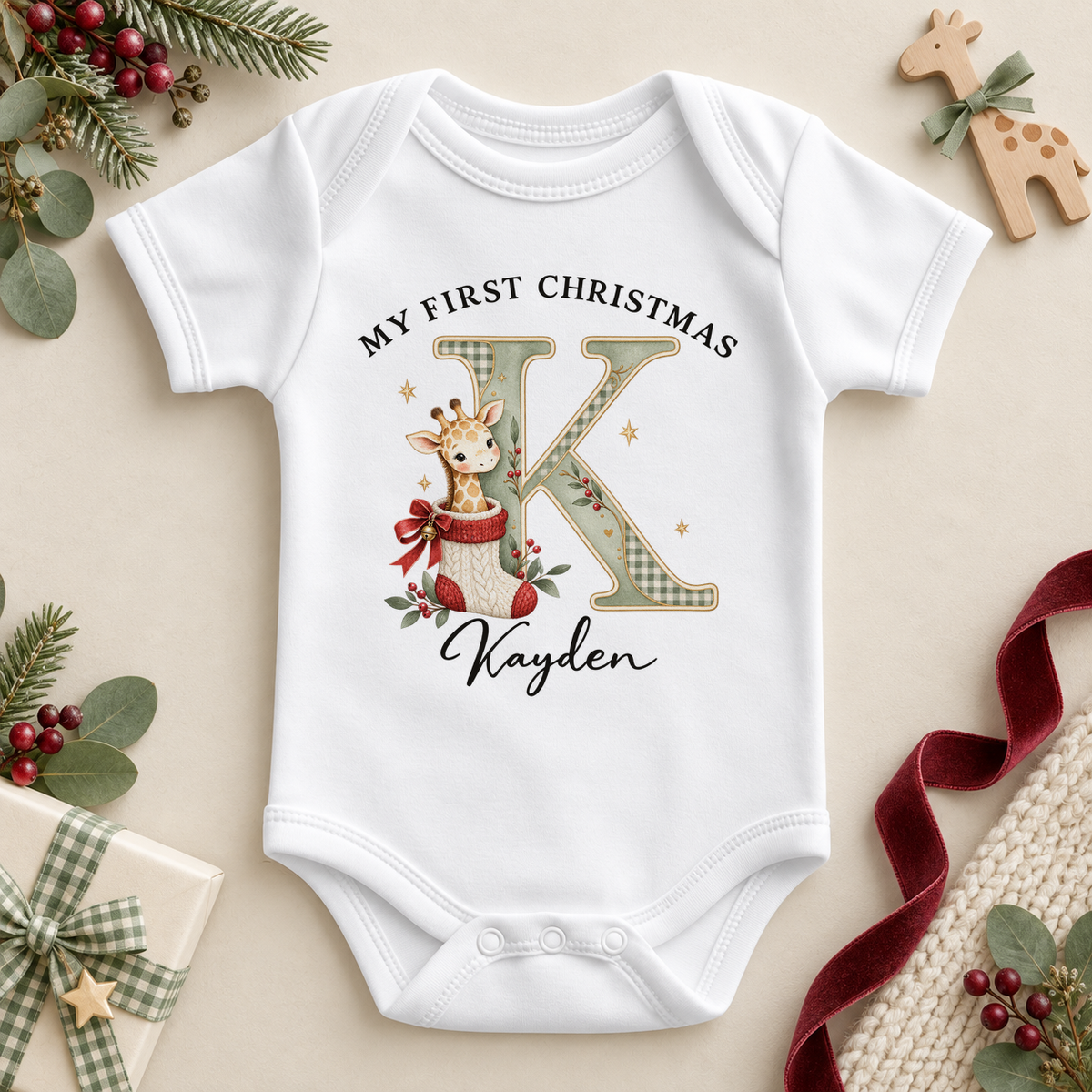 White baby bodysuit. The print features a cream and sage initial beside a giraffe peeking from a knitted Christmas stocking.