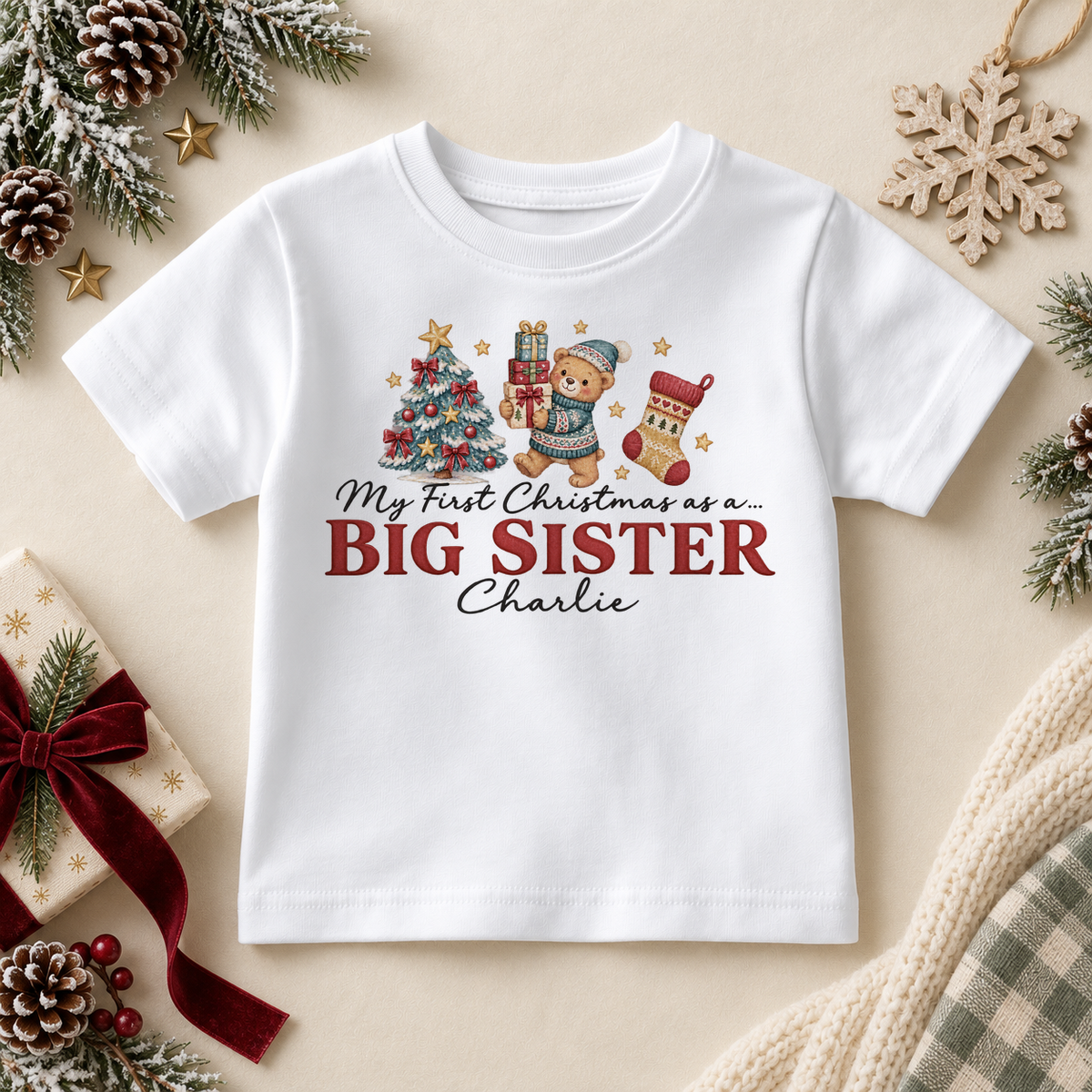 White children’s T-shirt. The print features a teddy holding gifts beside a tree and stocking above Big Sister lettering.