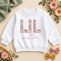 Lil Sister white sweatshirt with colourful floral LIL lettering and pink sister script, styled with flowers and sage fabric.