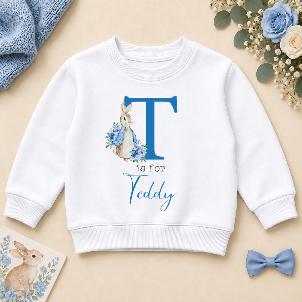 White personalised kids sweatshirt with a blue T initial, floral bunny in a blue jacket and T is for Teddy example name, styled on a cream surface.