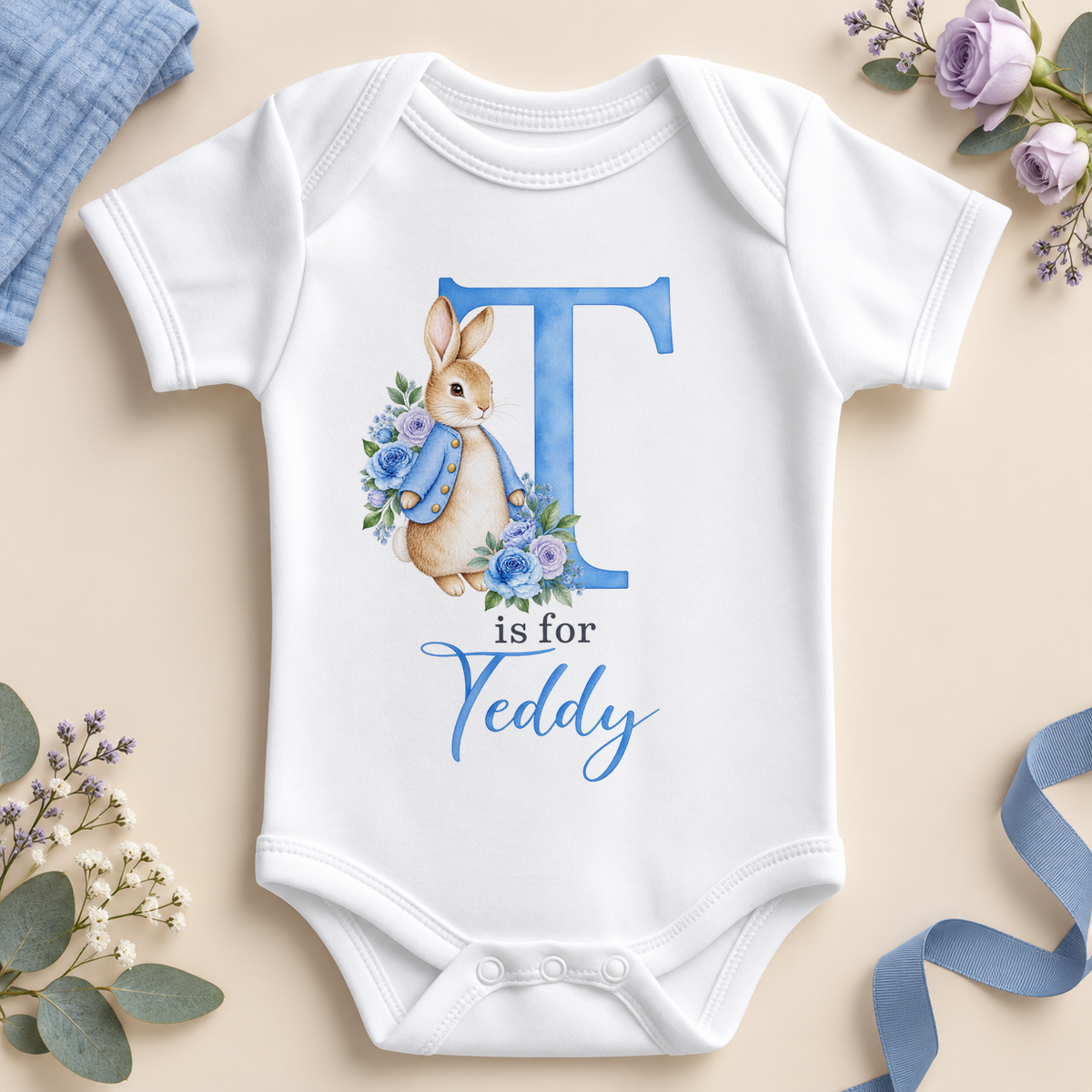 White personalised baby bodysuit with a large blue T initial, a watercolour bunny in a blue jacket with blue and lilac roses and T is for Teddy as an example name