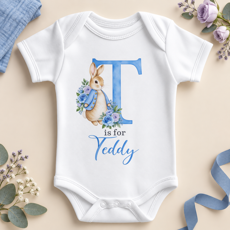 White personalised baby bodysuit with a large blue T initial, a watercolour bunny in a blue jacket with blue and lilac roses and T is for Teddy as an example name