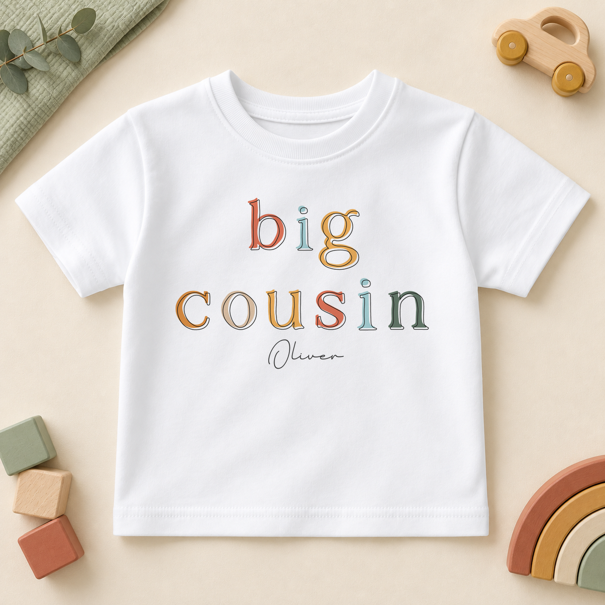 Personalised Big Cousin white kids T-shirt, colourful outlined serif letters with Oliver example name, styled with a wooden car, blocks and rainbow.