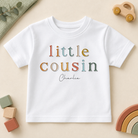 Personalised Little Cousin white kids T-shirt, colourful outlined serif letters with Charlie example name, styled with a wooden car, blocks and rainbow.