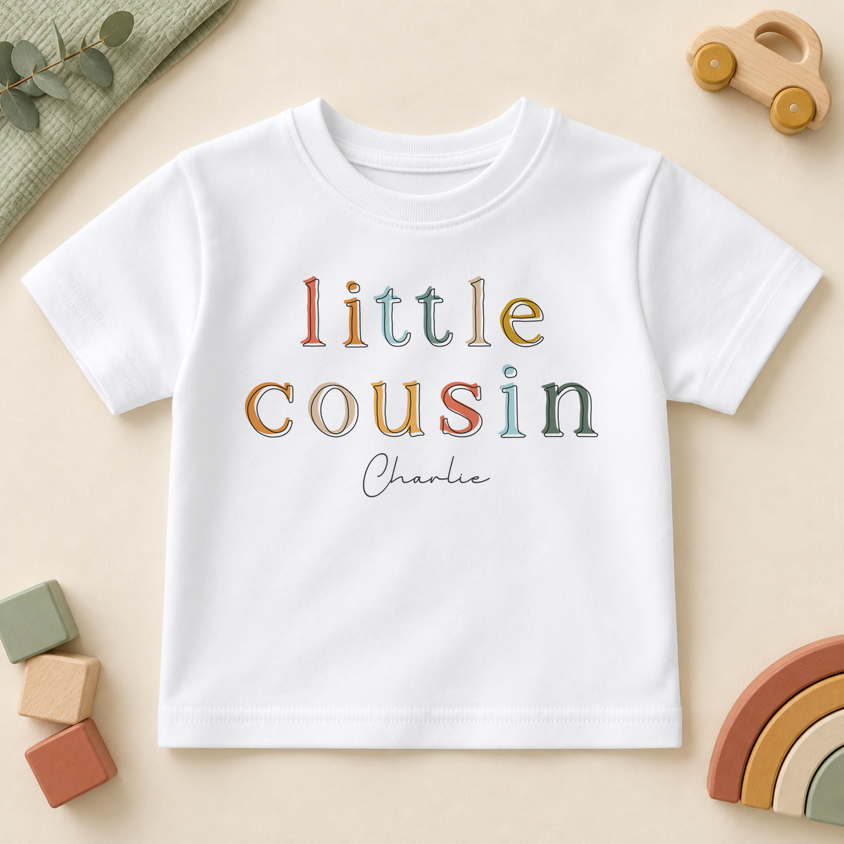 Personalised Little Cousin white kids T-shirt, colourful outlined serif letters with Charlie example name, styled with a wooden car, blocks and rainbow.