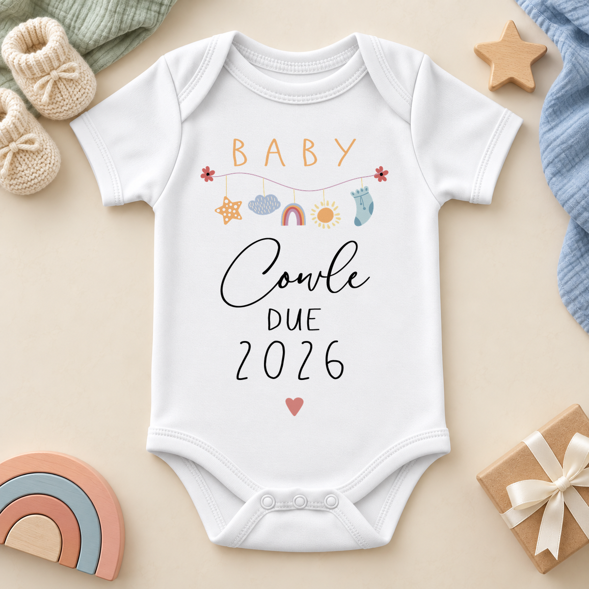 White baby announcement bodysuit with pastel hanging star, cloud, rainbow, sun and sock charms, centred BABY lettering, Cowle example surname and DUE2026, styled on a cream flat lay.
