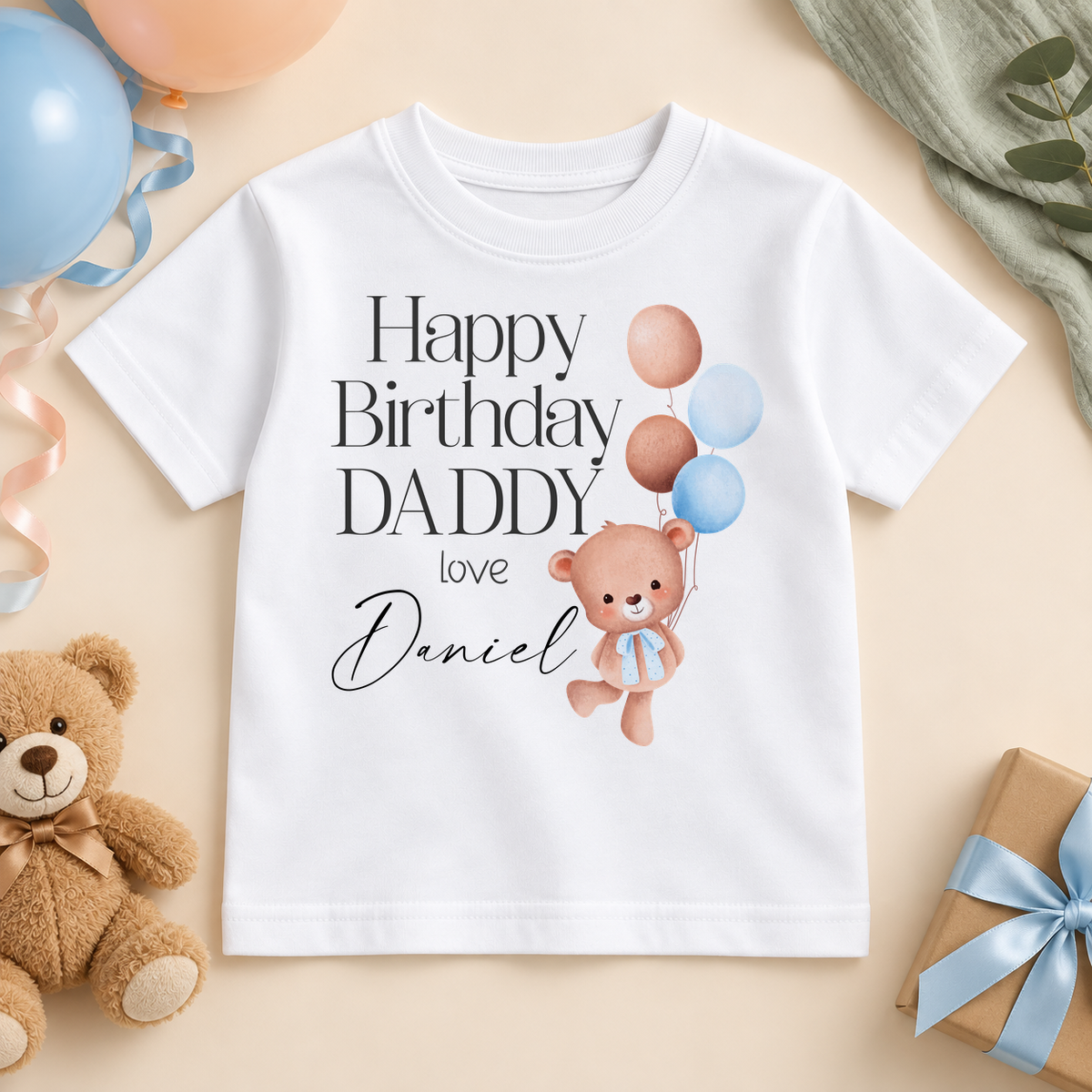Personalised white kids T-shirt with Happy Birthday DADDY, love Daniel example name and a teddy bear holding blue and peach balloons, styled with birthday balloons, a teddy and a gift.