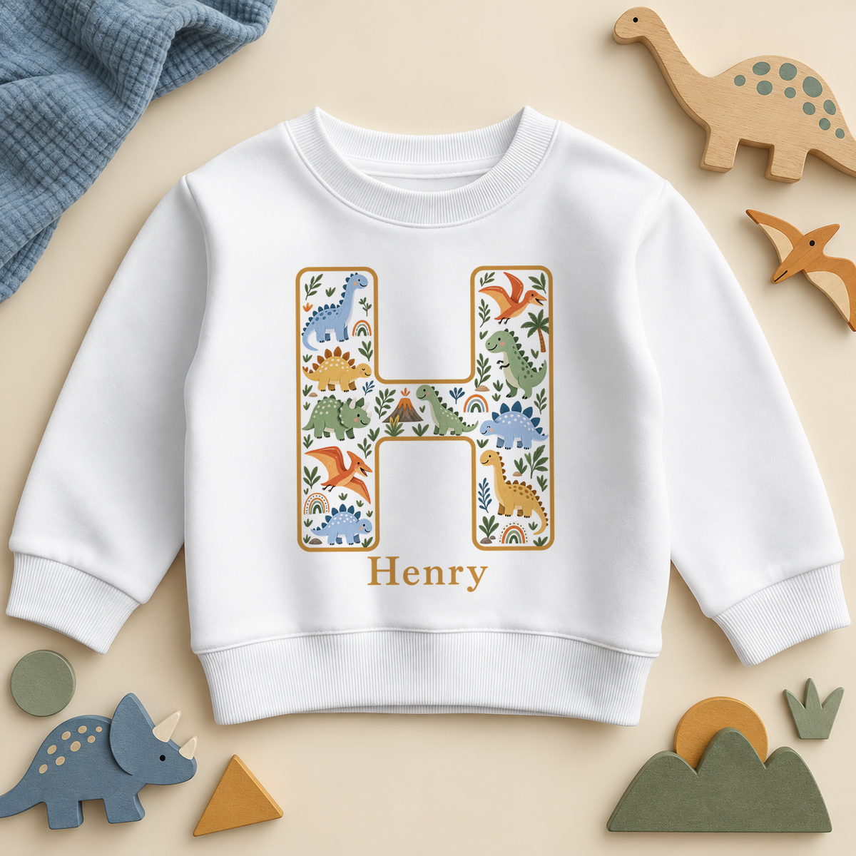 White kids sweatshirt with a mustard outlined initial H full of friendly dino characters, a little volcano and rainbows, with the example name Henry in mustard.