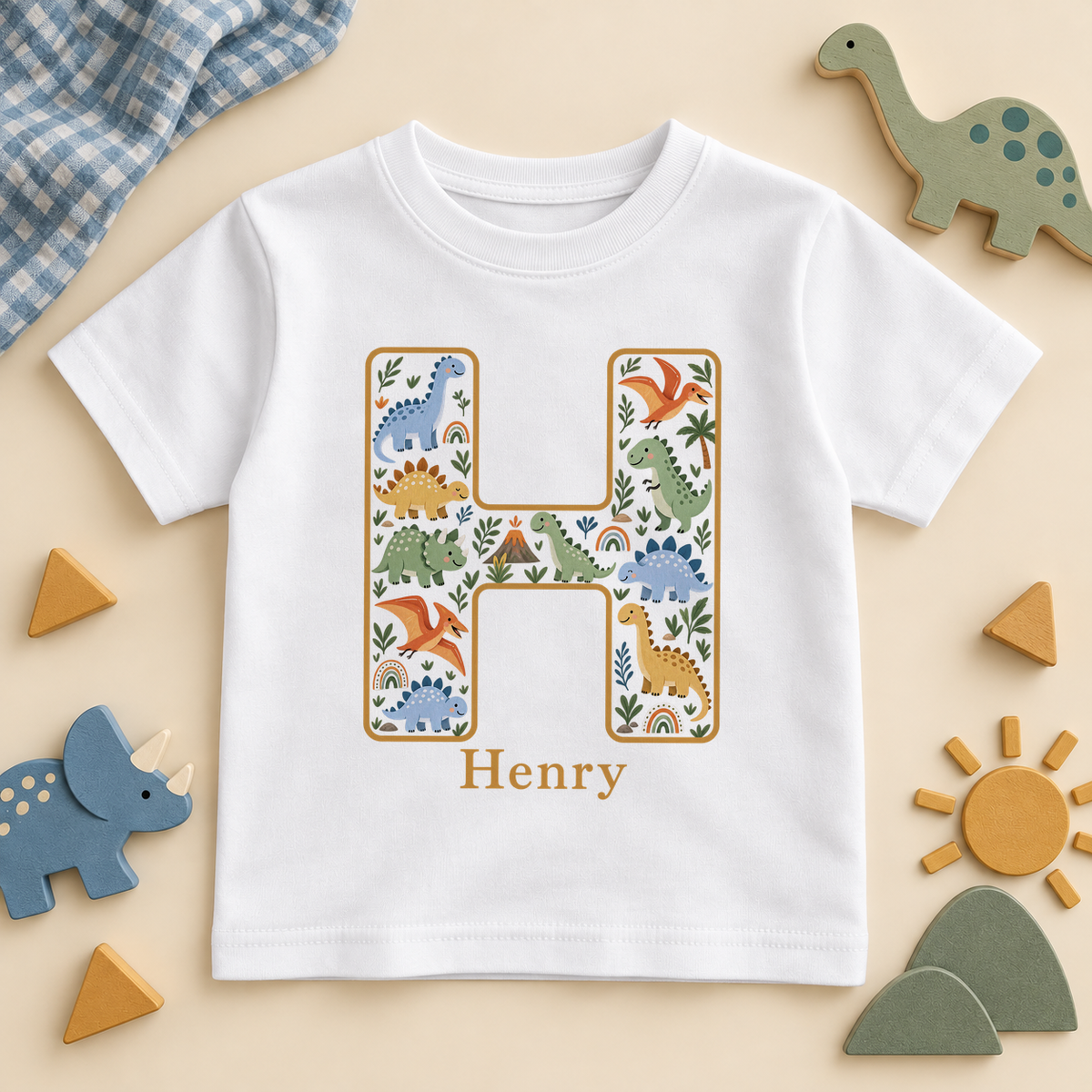 White kids T-shirt with a mustard outlined initial H full of friendly dino characters, a little volcano and rainbows, with the example name Henry in mustard.