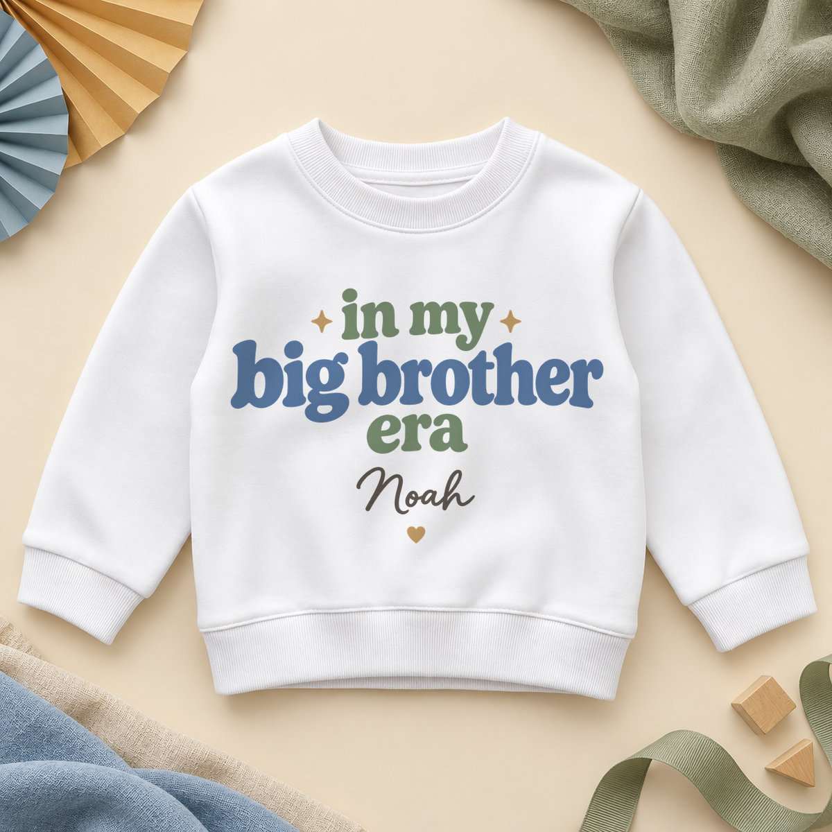 In my big brother era personalised kids sweatshirt: a white sweatshirt with retro blue and sage green lettering, a name in black script and a small gold heart