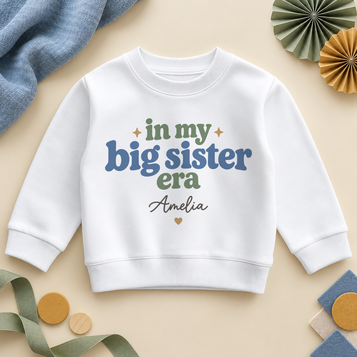 In my big sister era personalised kids sweatshirt: a white sweatshirt with retro blue and sage green lettering, a name in black script and a small gold heart