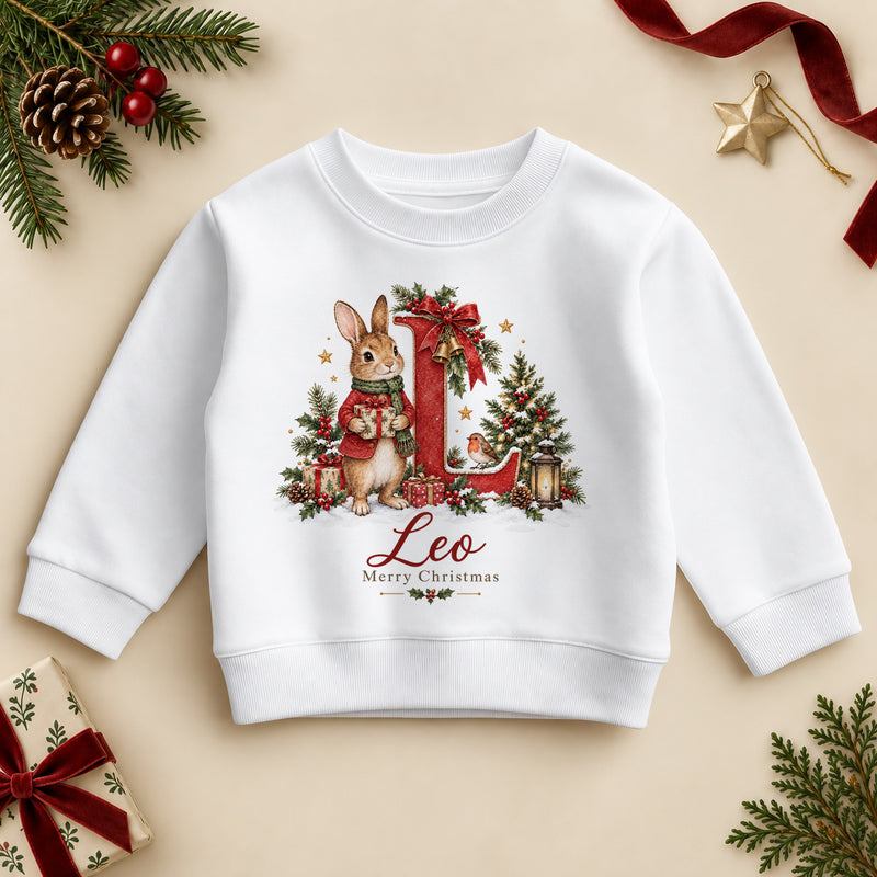 White kids' sweatshirt with a storybook bunny rabbit in a red jacket holding a present beside a large red initial, holly, a robin and a Christmas tree, a name in red script and Merry Christmas. Example name shown.
