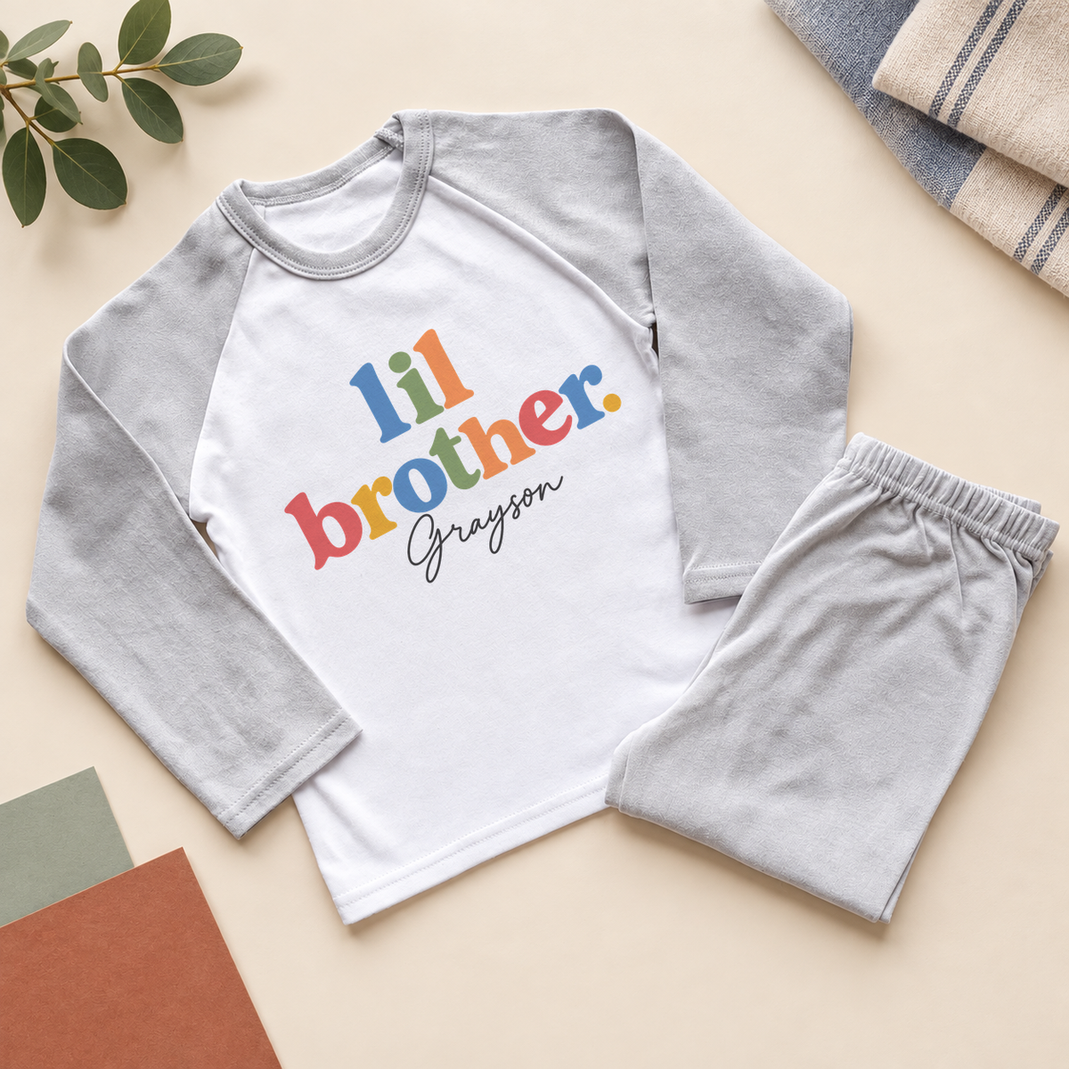 Personalised lil brother pyjamas: "lil brother." in colourful lowercase letters with a name in dark script on a white chest, light grey raglan sleeves and grey trousers