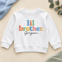Personalised lil brother sweatshirt with lil brother in chunky pastel rainbow letters and an example name in black script on a white crew neck sweatshirt