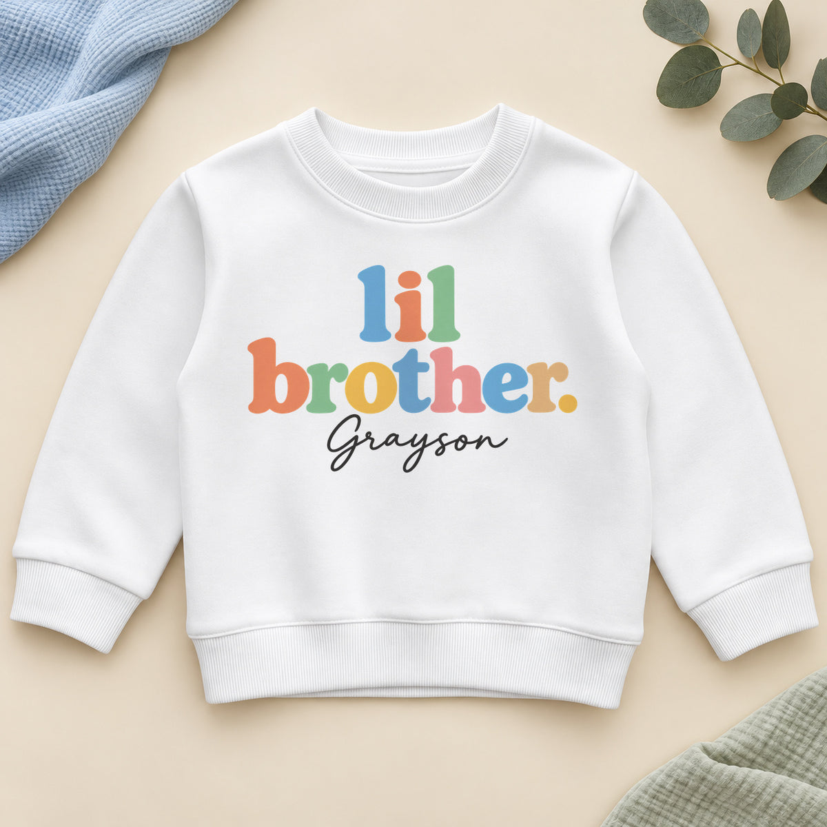 Personalised lil brother sweatshirt with lil brother in chunky pastel rainbow letters and an example name in black script on a white crew neck sweatshirt