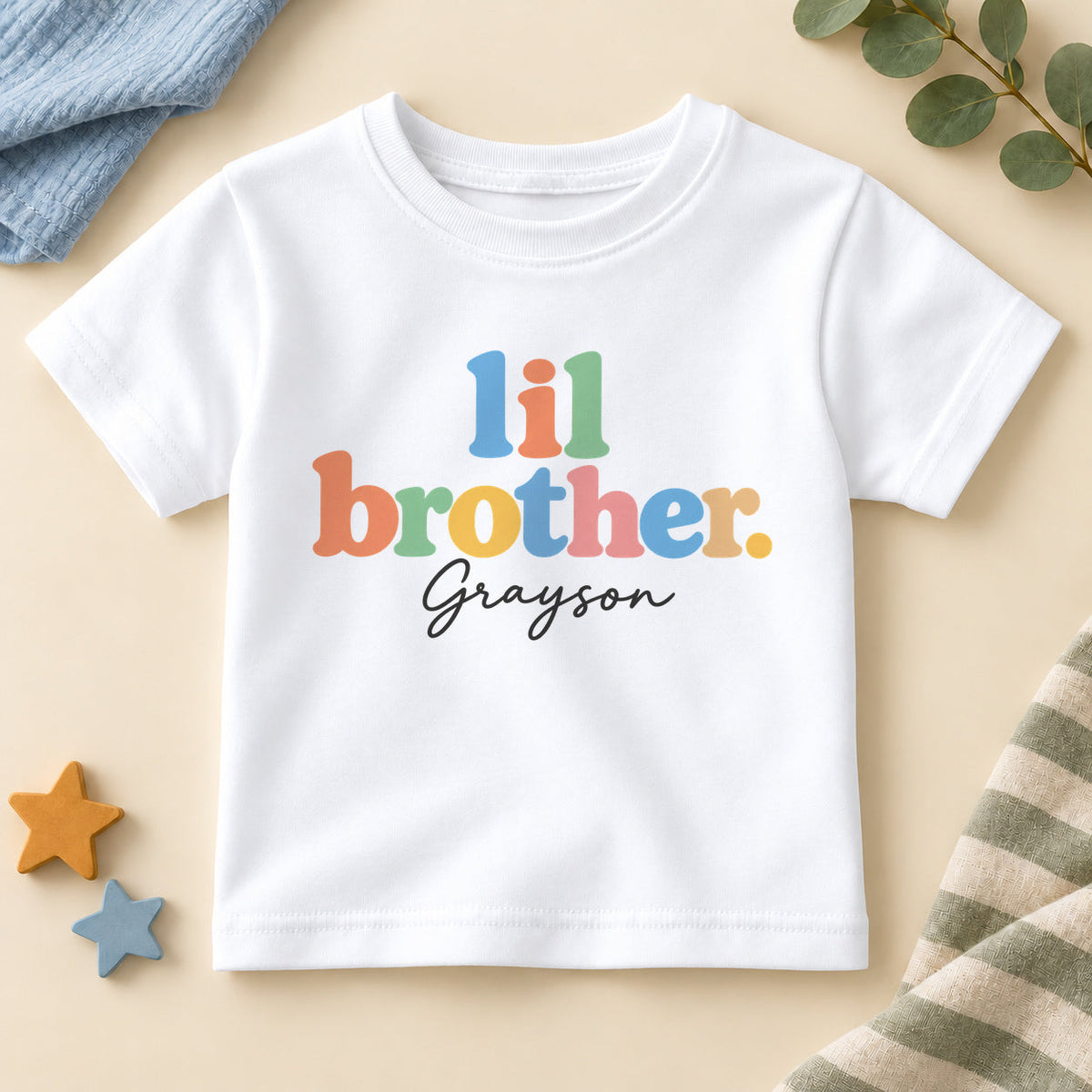 Personalised lil brother T-shirt with lil brother in chunky pastel rainbow letters and an example name in black script on a white crew neck tee