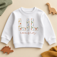 Lil cousin dino sweatshirt with LIL in capital letters filled with a cartoon dinosaur and rainbow print and cousin in terracotta script on a white crew neck sweatshirt