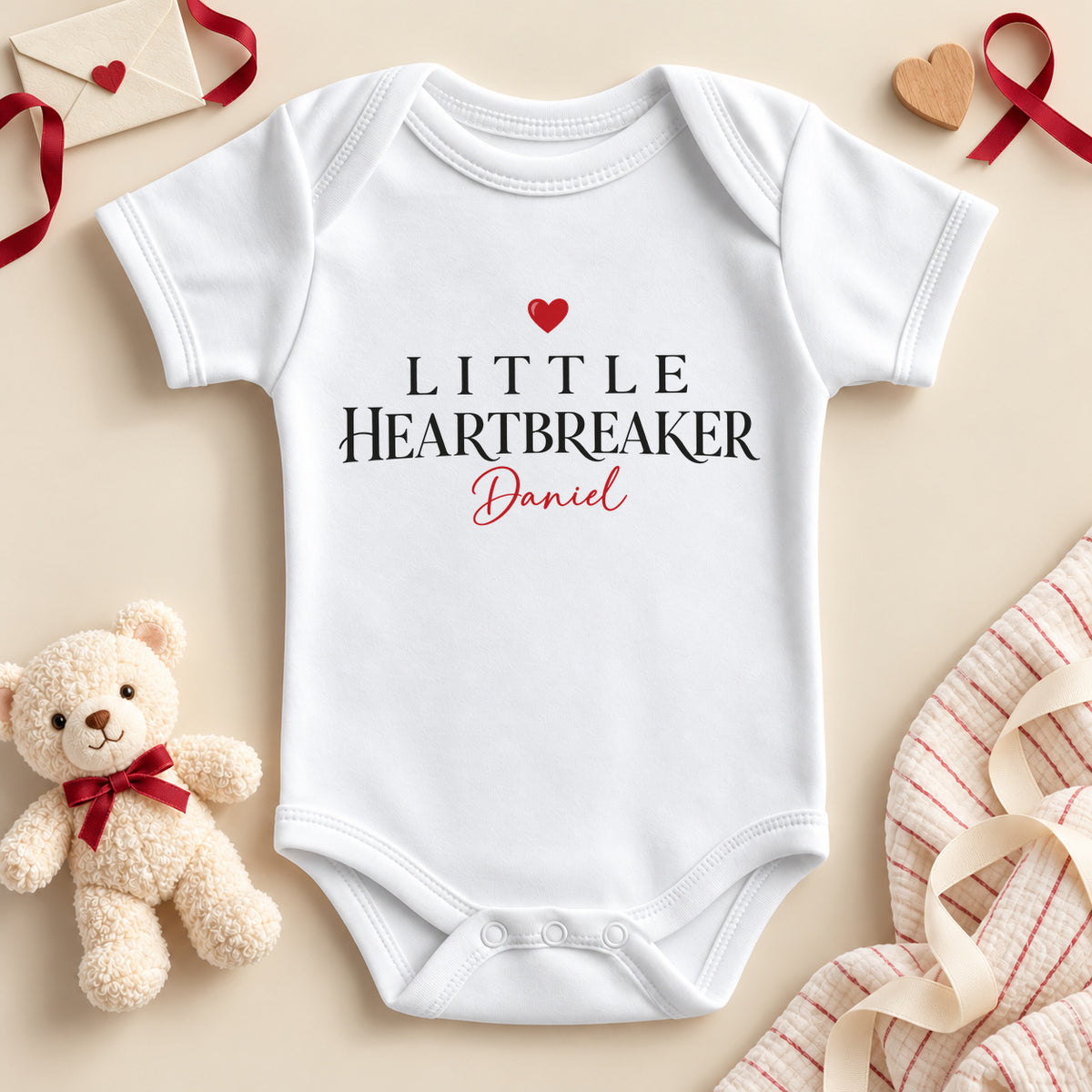 Personalised Little Heartbreaker baby bodysuit: a white baby vest with black serif capitals, a red heart and an example name in red script