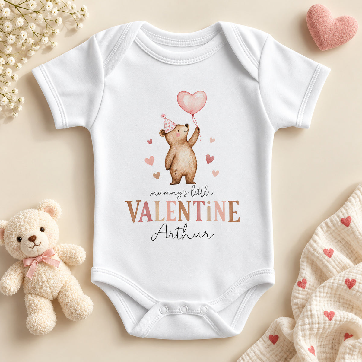 Personalised Mummy's little valentine baby bodysuit: a white baby vest with a teddy bear in a pink party hat holding a heart balloon, brown and pink lettering and an example name