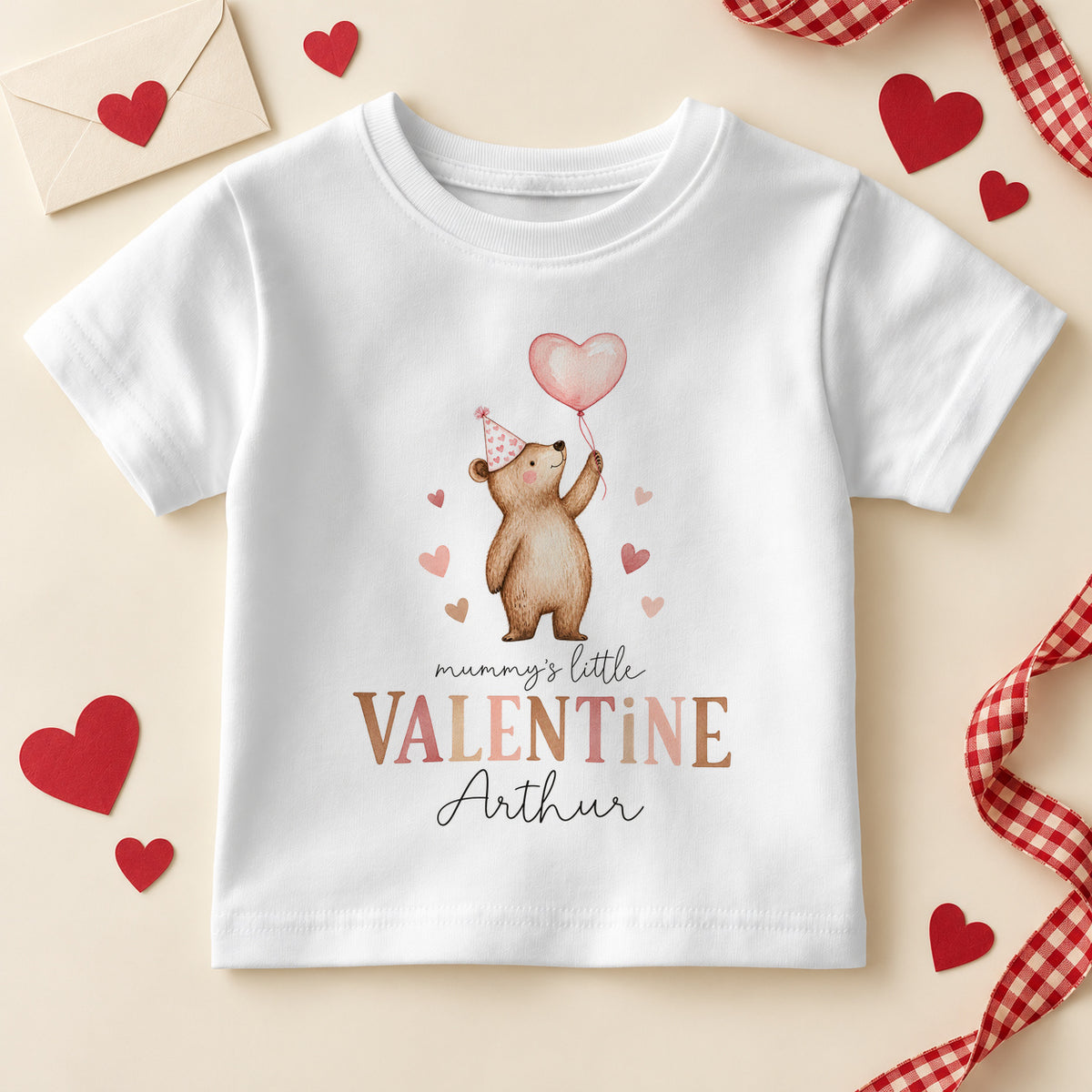 Personalised Mummy's little valentine kids T-shirt: a white tee with a teddy bear in a pink party hat holding a heart balloon, brown and pink lettering and an example name