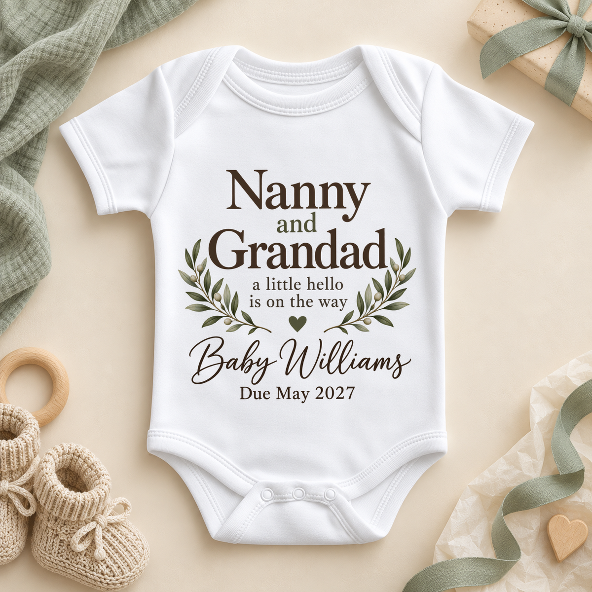 Nanny and Grandad pregnancy announcement baby vest bodysuit: a white baby bodysuit with brown lettering, olive leaf branches, a green heart, a surname and a due date