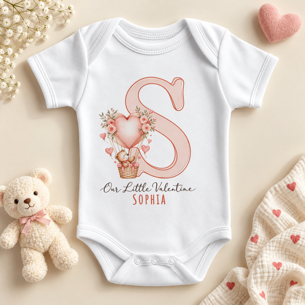 Personalised Our Little Valentine baby bodysuit: a white baby vest with a large pink initial, a teddy bear in a heart balloon with roses and an example name