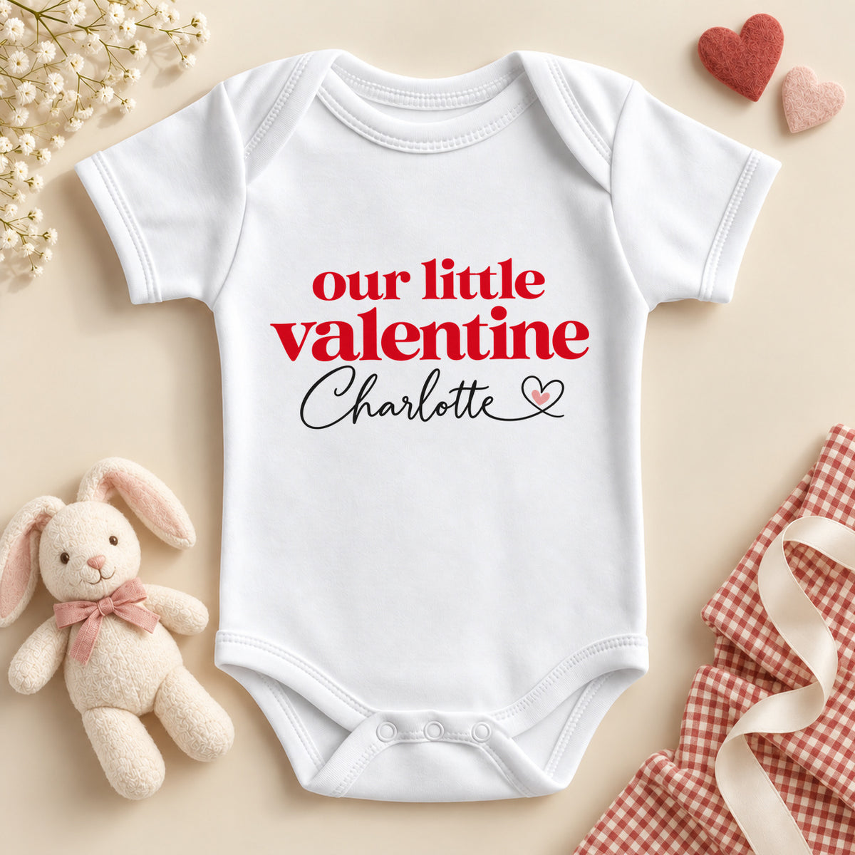 Personalised Our Little Valentine baby bodysuit: a white baby vest with bold red lettering and an example name in black script with a heart flourish