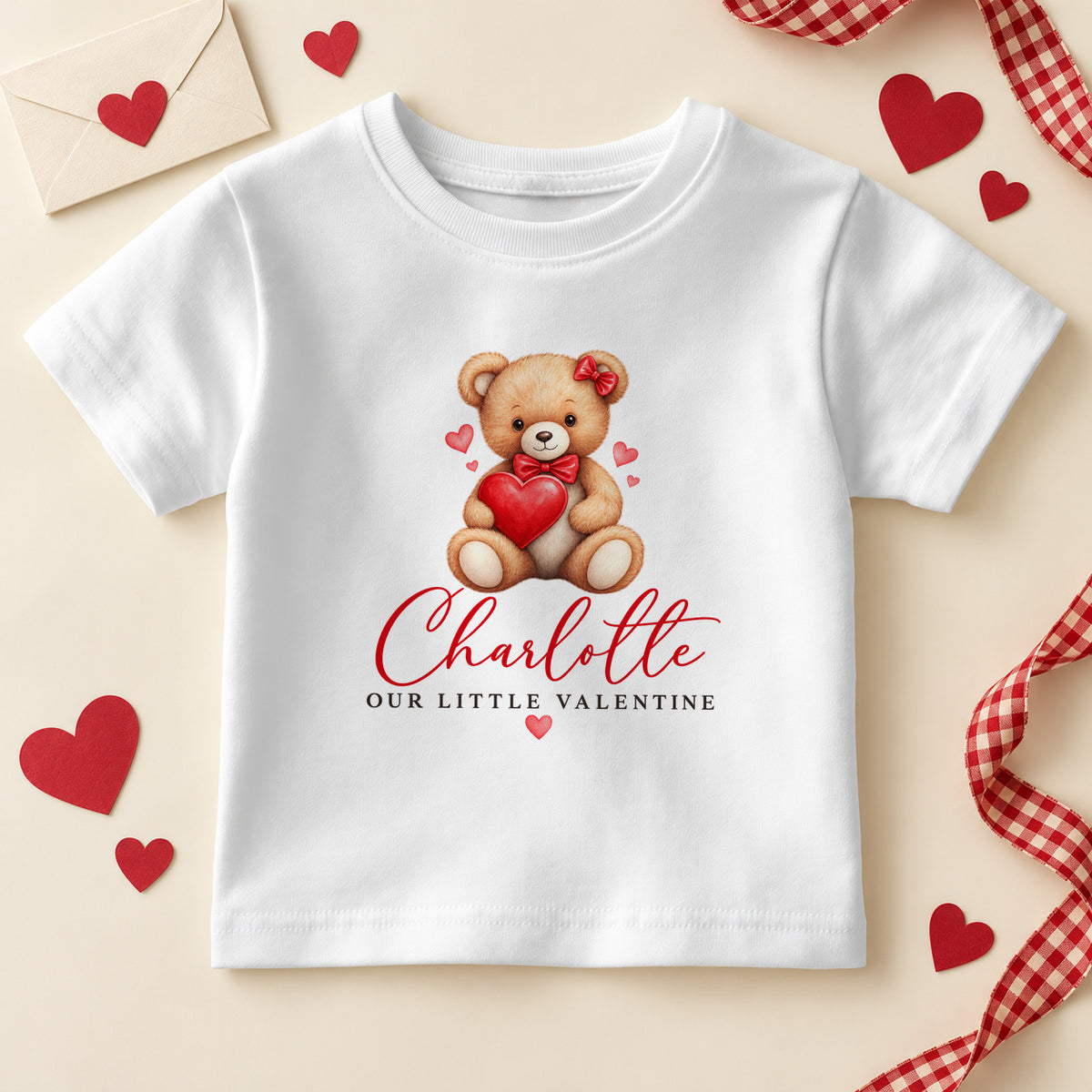 Personalised Our Little Valentine kids T-shirt: a white tee with a teddy bear in a red bow hugging a big red heart and an example name in red script